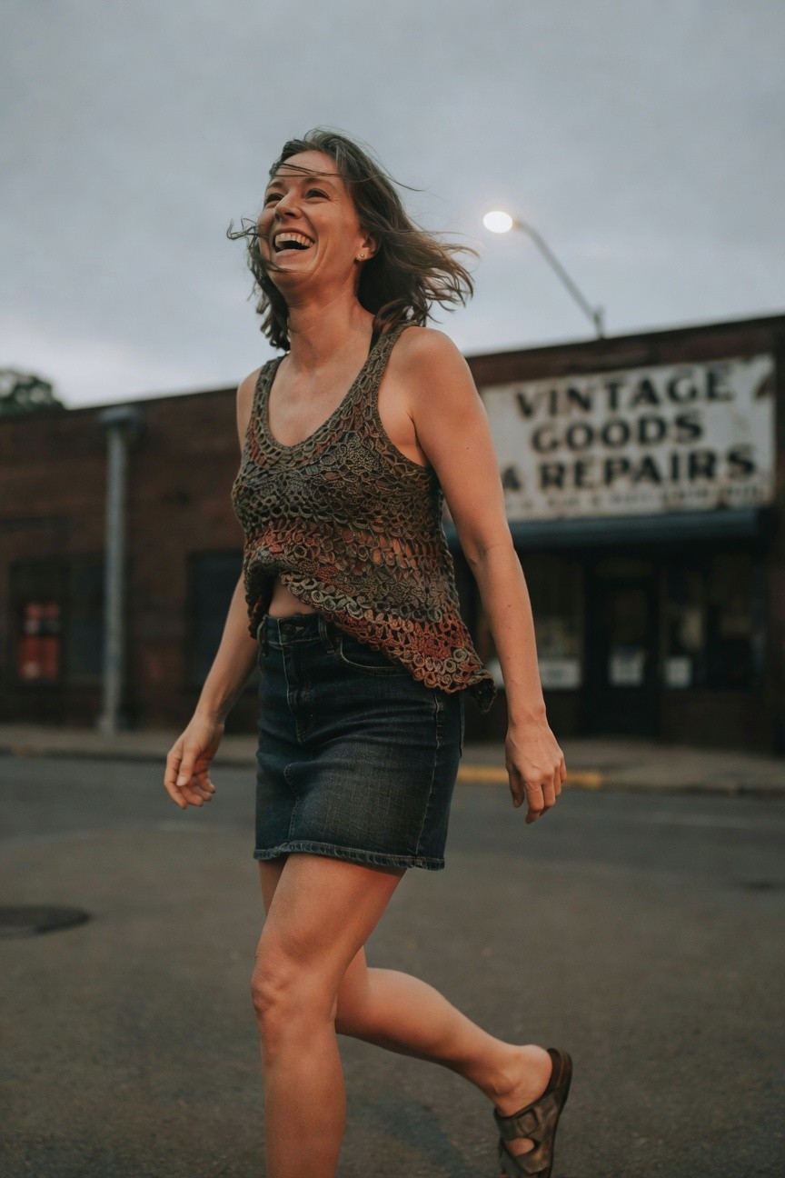 Woman walking in open-knit multicolored tank top with rust, olive, and brown tones over a low-rise short denim skirt and flat woven sandals, midriff slightly exposed, joyful expression
