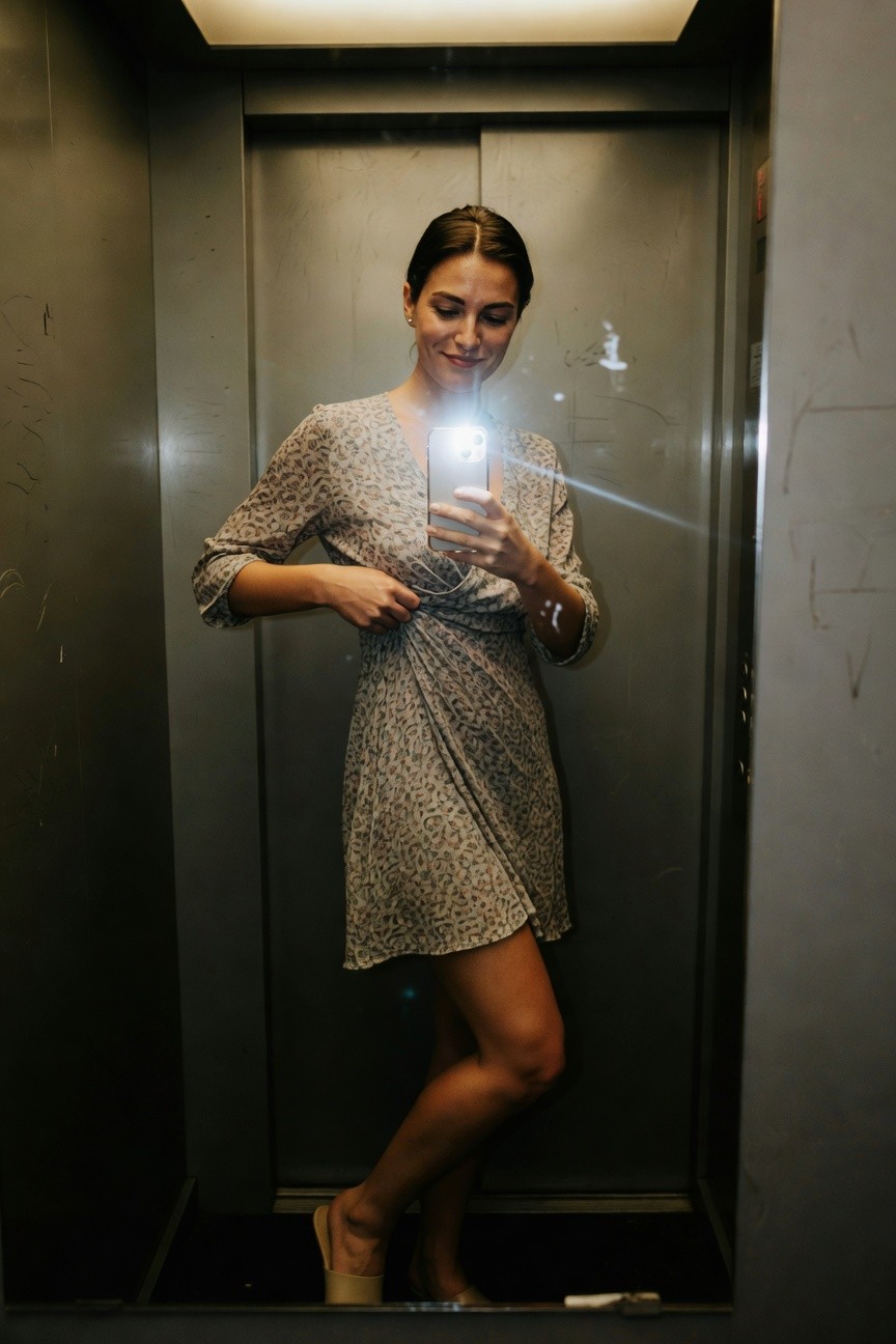 Woman in elevator mirror selfie wearing a light gray-toned leopard print wrap dress with long sleeves and mid-thigh hem, nude slide sandals, holding phone for photo