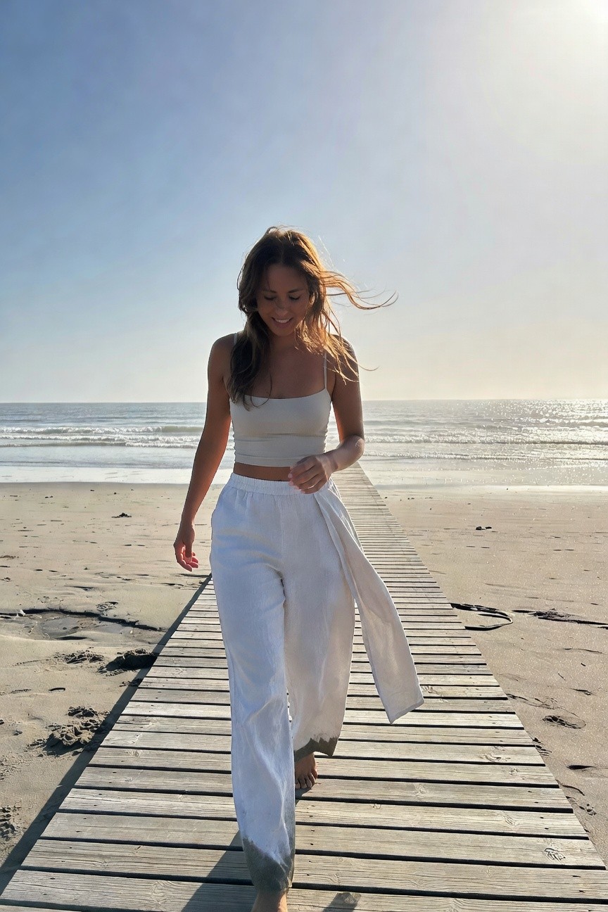 Woman in beige strapless crop top and flowing white wide-leg pants walking barefoot on wooden boardwalk, hair blowing in wind, ocean visible in background
