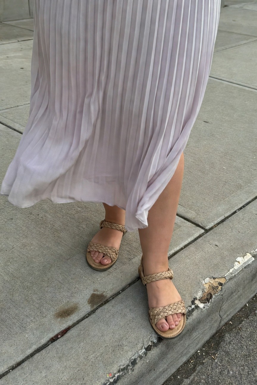 Close-up of a plus-size woman's lower outfit featuring a pale lavender pleated midi skirt flowing over tan braided strap sandals on a concrete sidewalk