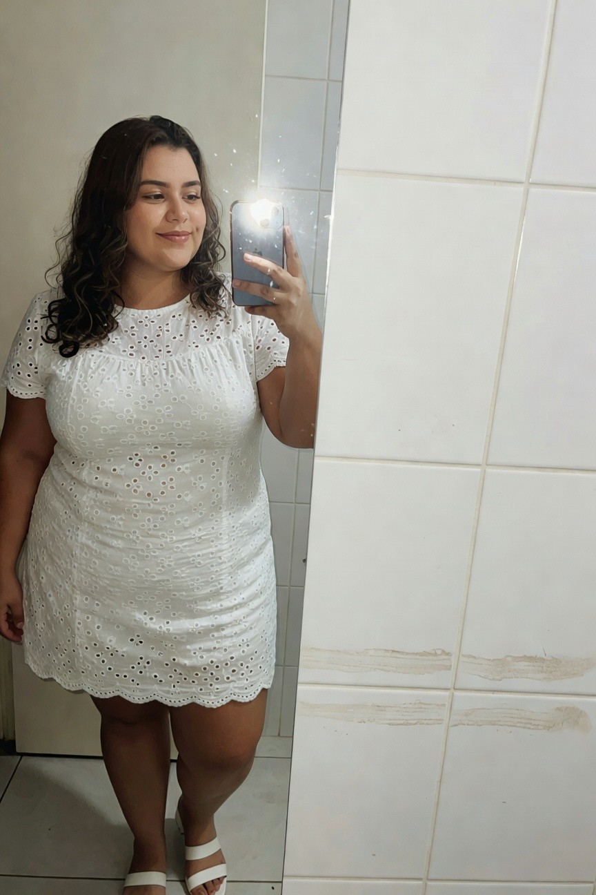 Plus-size woman in a short white eyelet cotton dress with short sleeves and scalloped hem, posing in front of a bathroom mirror while holding her phone for a selfie, wearing white strappy flat sandals