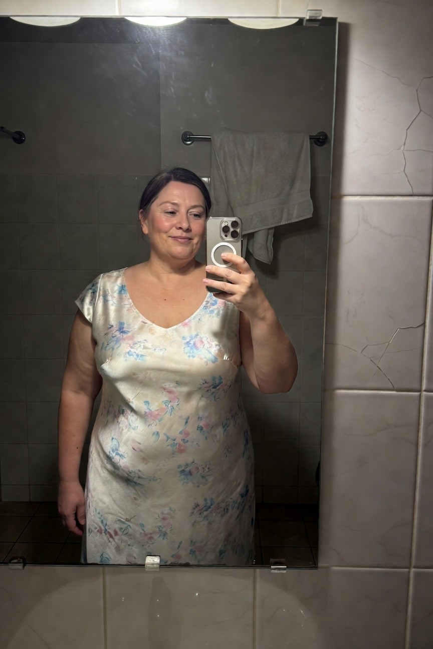 Plus-size woman smiling in a selfie wearing a light-colored sleeveless silk dress with floral print in a bathroom mirror, towel on rack nearby