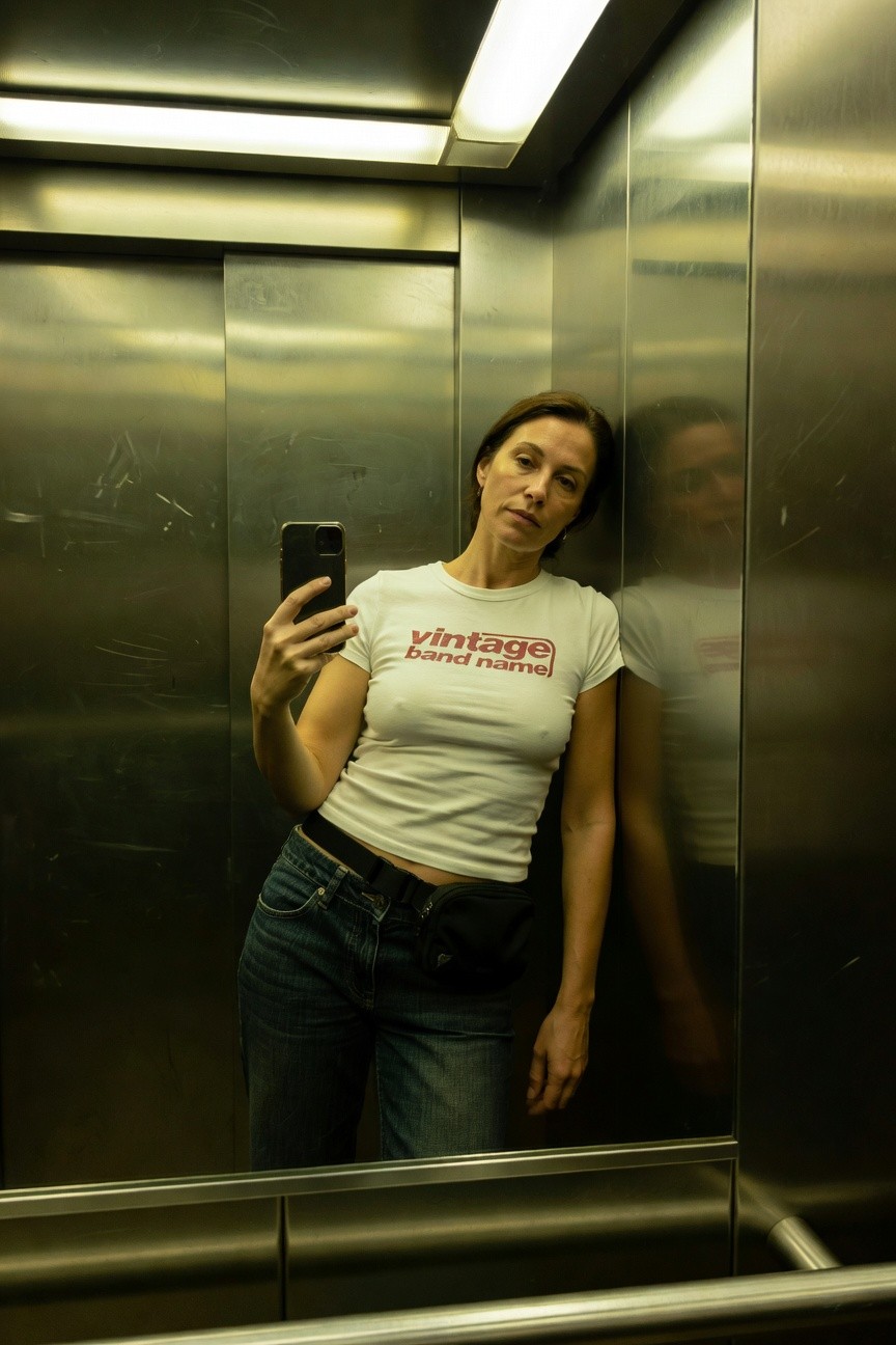 Woman taking a mirror selfie in an elevator, wearing a fitted white T-shirt with red "Vintage Bandname" text, low-rise baggy blue jeans, and a black fanny pack buckled at the waist, leaning casually against the wall