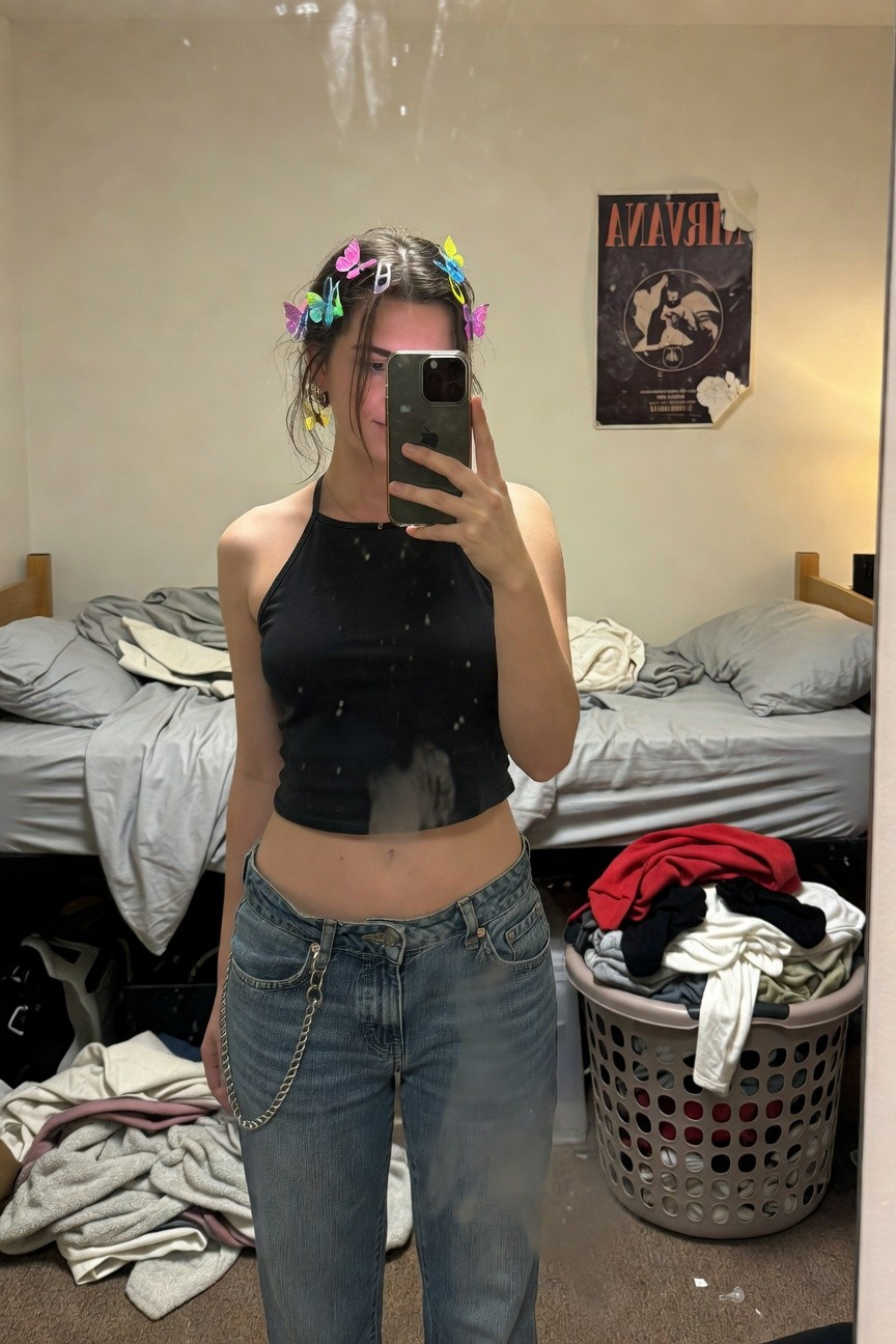Mirror selfie of a woman in a black strappy cropped halter top exposing midriff, light blue baggy low-rise jeans with silver chain detail, colorful butterfly hair clips in wavy brown hair, gold hoop earrings, messy dorm room with laundry and Nirvana poster in background