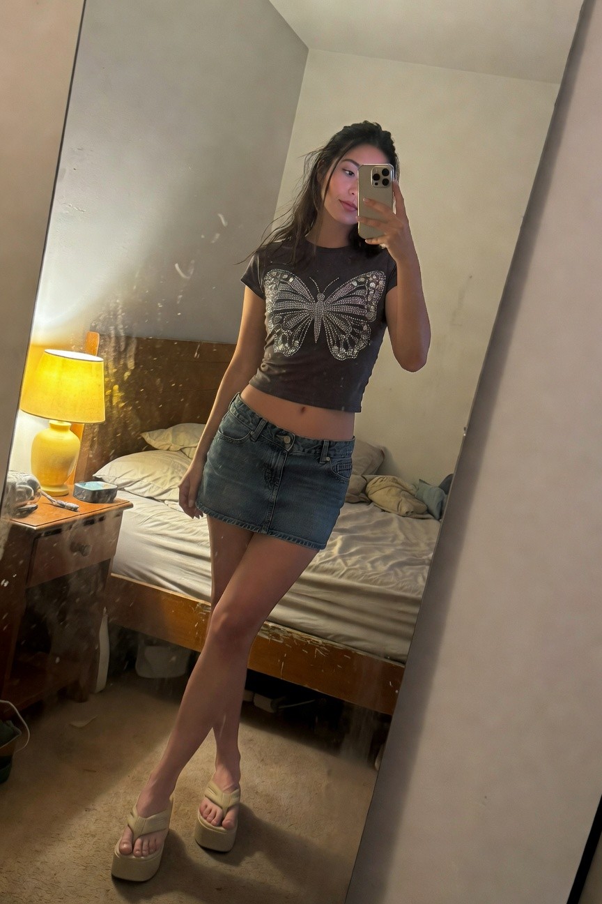 Mirror selfie of woman in dark gray cropped t-shirt with large silver-embellished butterfly design, low-rise faded blue denim mini skirt exposing midriff, and beige platform flip-flop sandals, posing confidently in bedroom setting