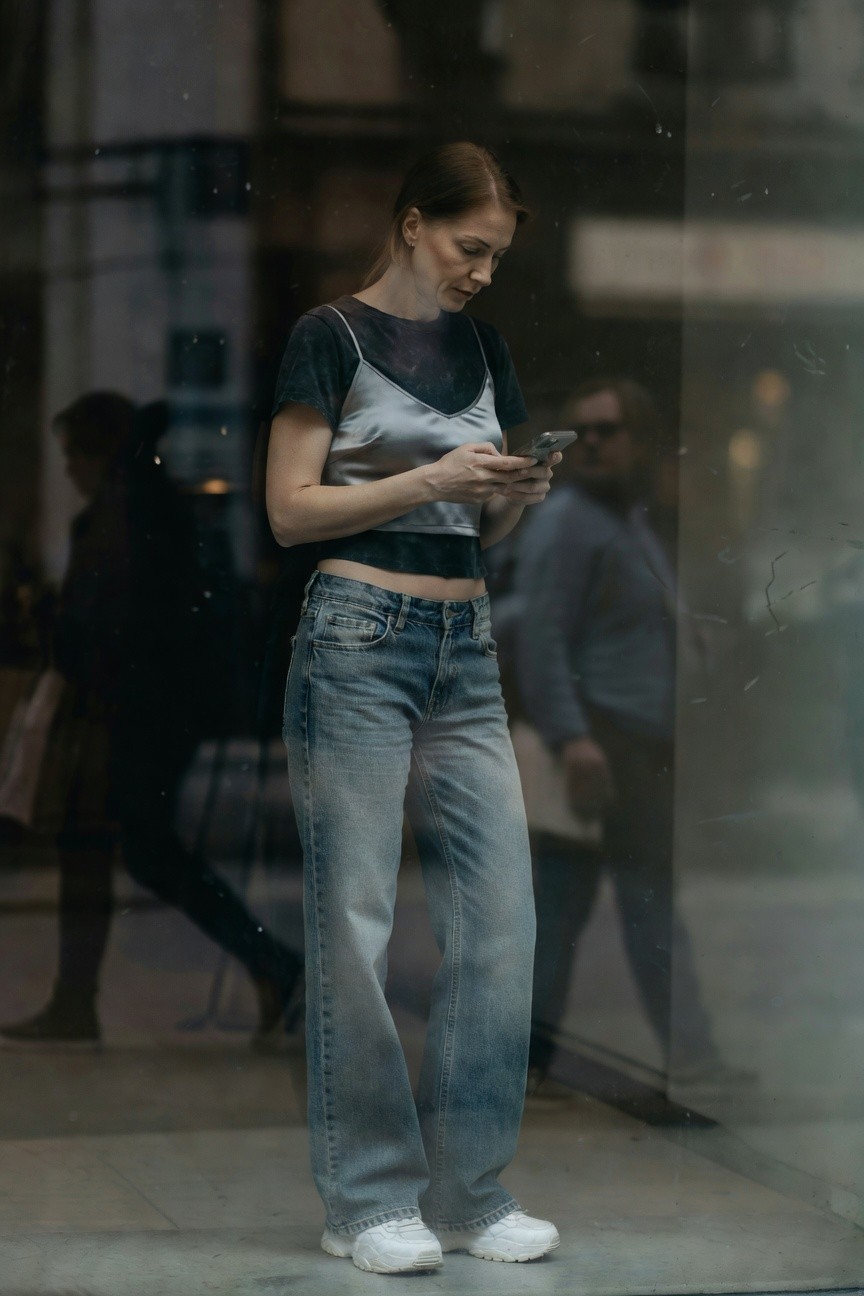 Woman standing in front of glass window wearing cropped black short-sleeve tee layered with shiny silver camisole, low-rise baggy light blue jeans, and white chunky sneakers, holding phone while viewing her reflection