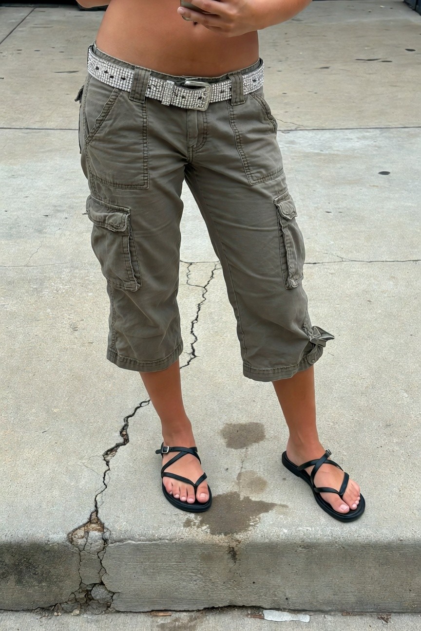 Young woman standing on cracked concrete wearing low-rise olive green cargo capri pants with multiple pockets, accented by a wide white rhinestone-studded belt low on her hips, paired with black cross-strap flat sandals exposing her toes, bare midriff visible above the pants