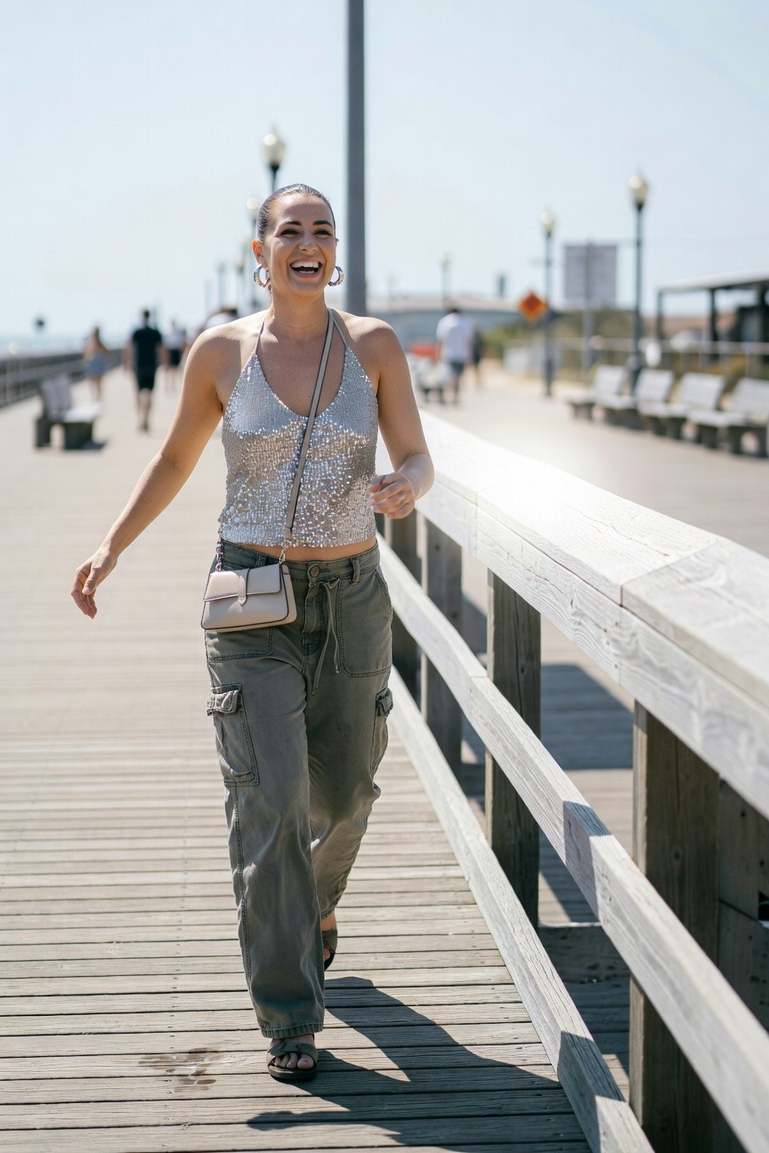 Smiling woman in silver sequin halter crop top, olive green cargo pants, beige crossbody bag, gold hoop earrings, and strappy sandals walking along a wooden pier railing