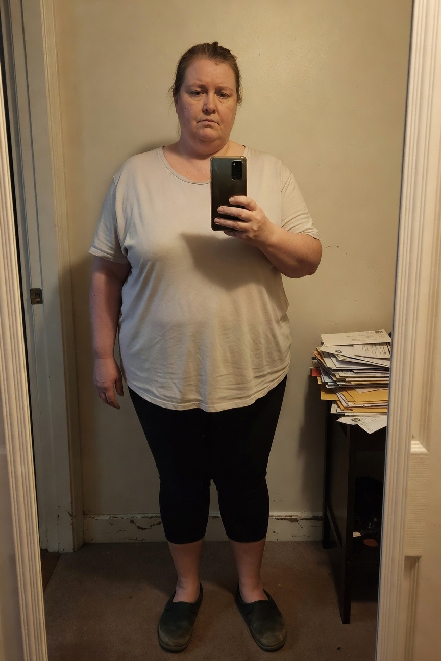 Plus-size woman in a loose light gray short-sleeve t-shirt tucked loosely over black capri-length leggings, wearing dark slip-on shoes, taking a mirror selfie in a hallway with stacks of mail nearby