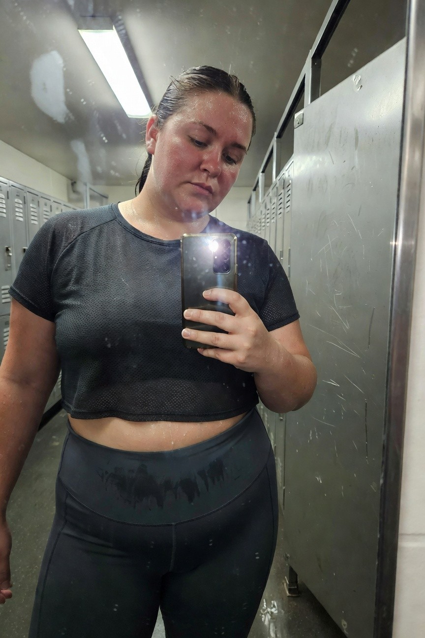 Plus-size woman taking a sweaty post-workout selfie in a locker room, wearing a black mesh cropped top that reveals midriff and high-waisted black leggings, phone in hand with gym lockers and mirrors in background