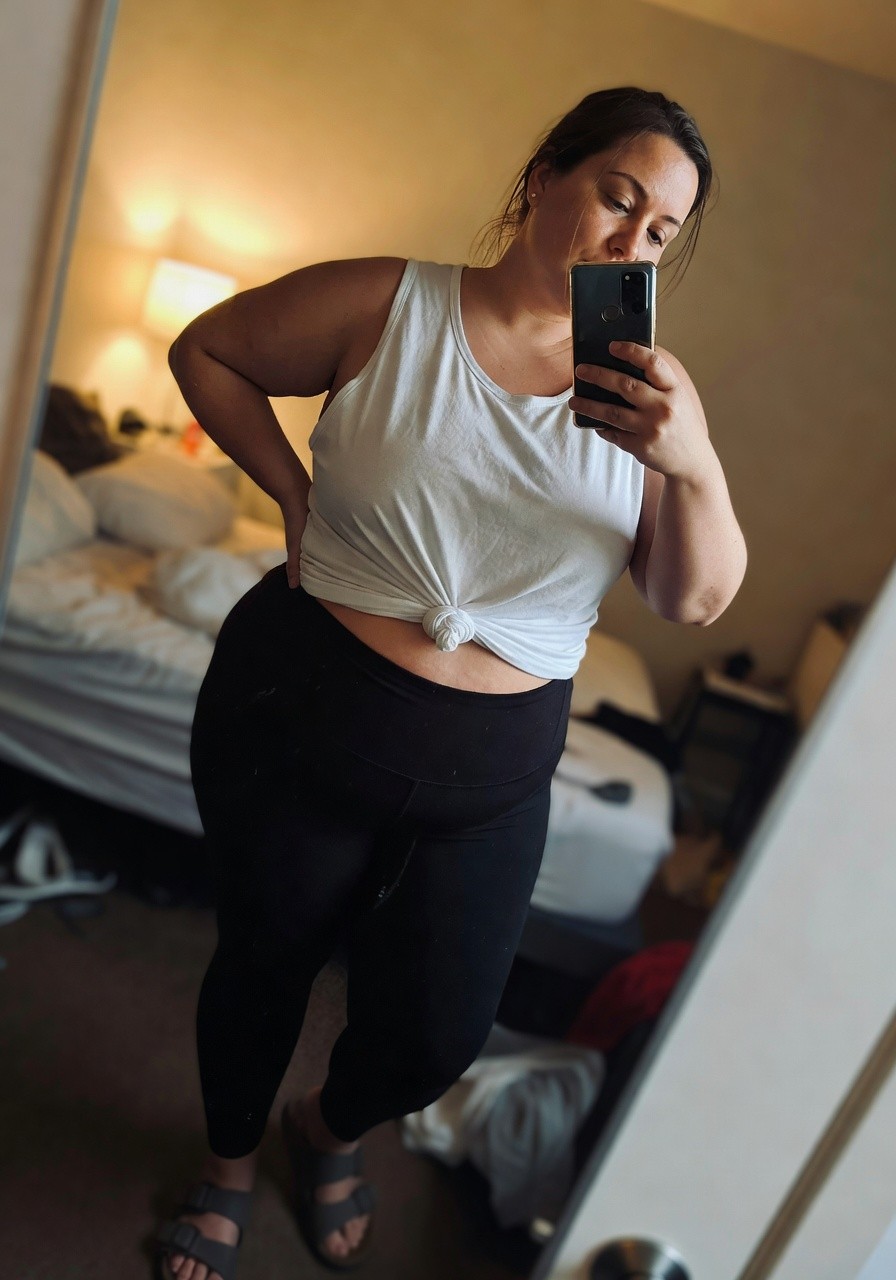 Plus-size woman in mirror selfie wearing a white sleeveless tank top knotted at the waist over black high-waisted leggings and brown sandals, standing confidently in a bedroom.
