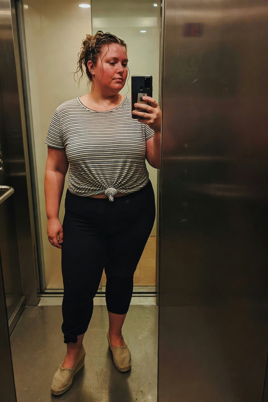 Plus-size woman taking elevator mirror selfie in light gray and white horizontal striped short-sleeve crop top tied at front, black capri-length leggings cuffed at ankles, and beige espadrille wedge sandals, hair in messy bun post-shower glow.