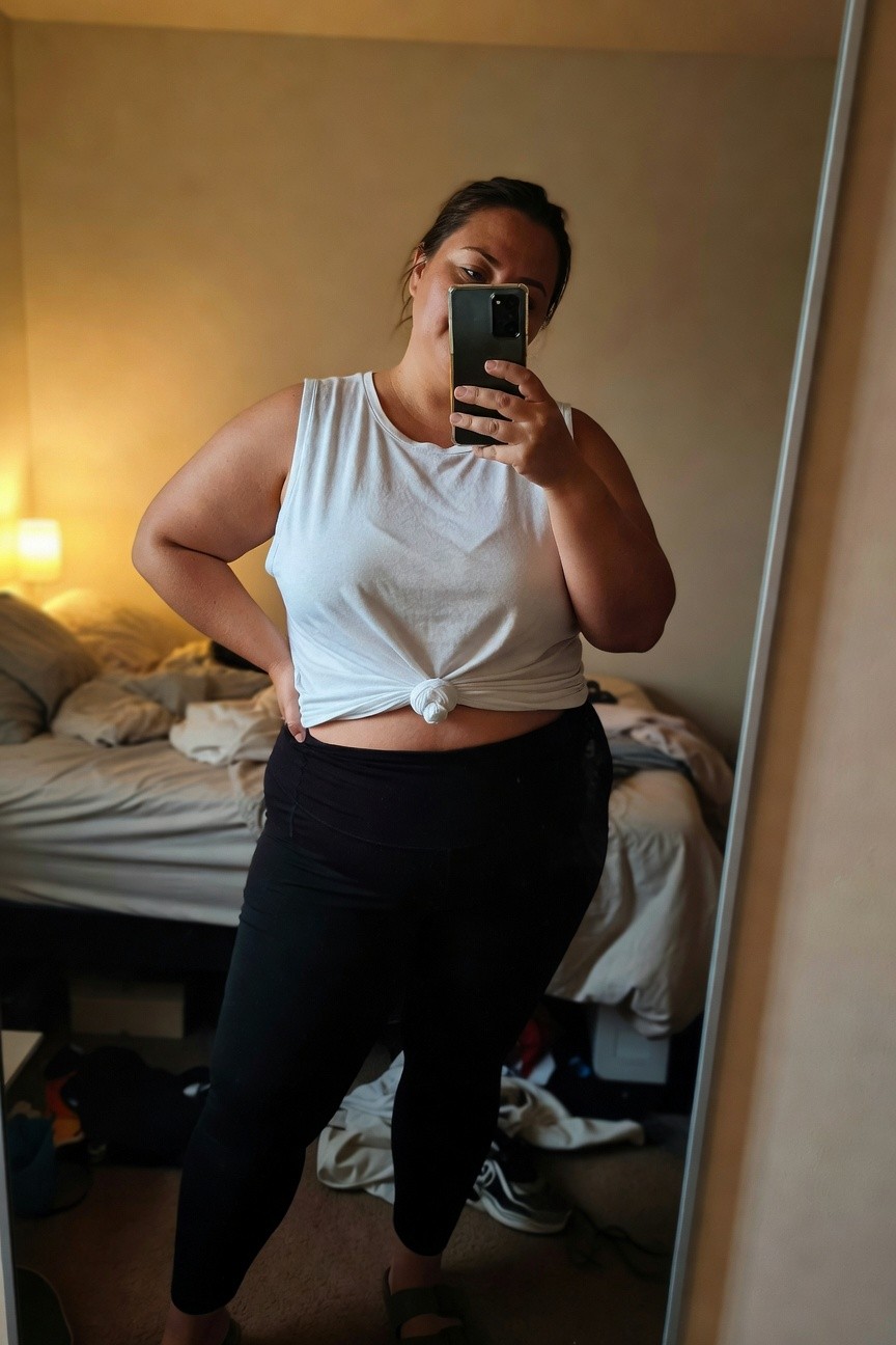 Plus-size woman in mirror selfie wearing a white sleeveless tank top knotted at the waist over black high-waisted leggings and brown sandals, standing confidently in a bedroom.