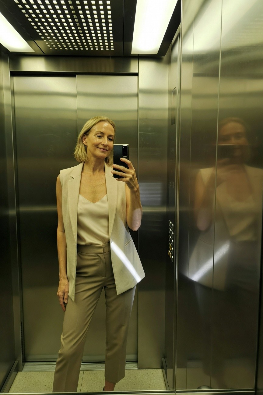 Mature blonde woman in a cream sleeveless vest over a silk camisole and matching beige wide-leg trousers, taking a mirror selfie in a metallic elevator with her reflection visible.