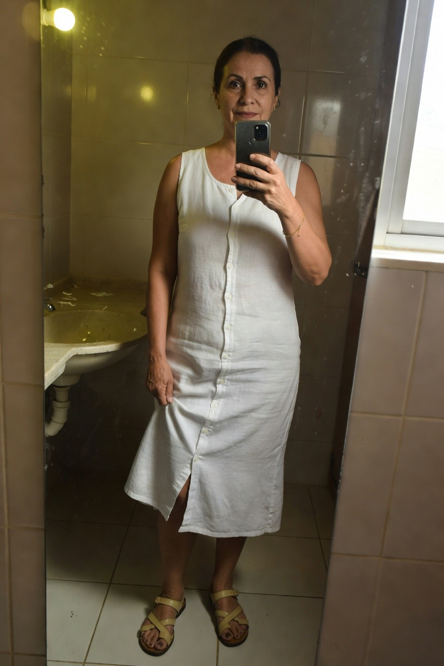 Mature woman in her 50s taking a mirror selfie in a bathroom, wearing a sleeveless white linen button-front midi dress with side slit, gold bracelet, and yellow strappy sandals