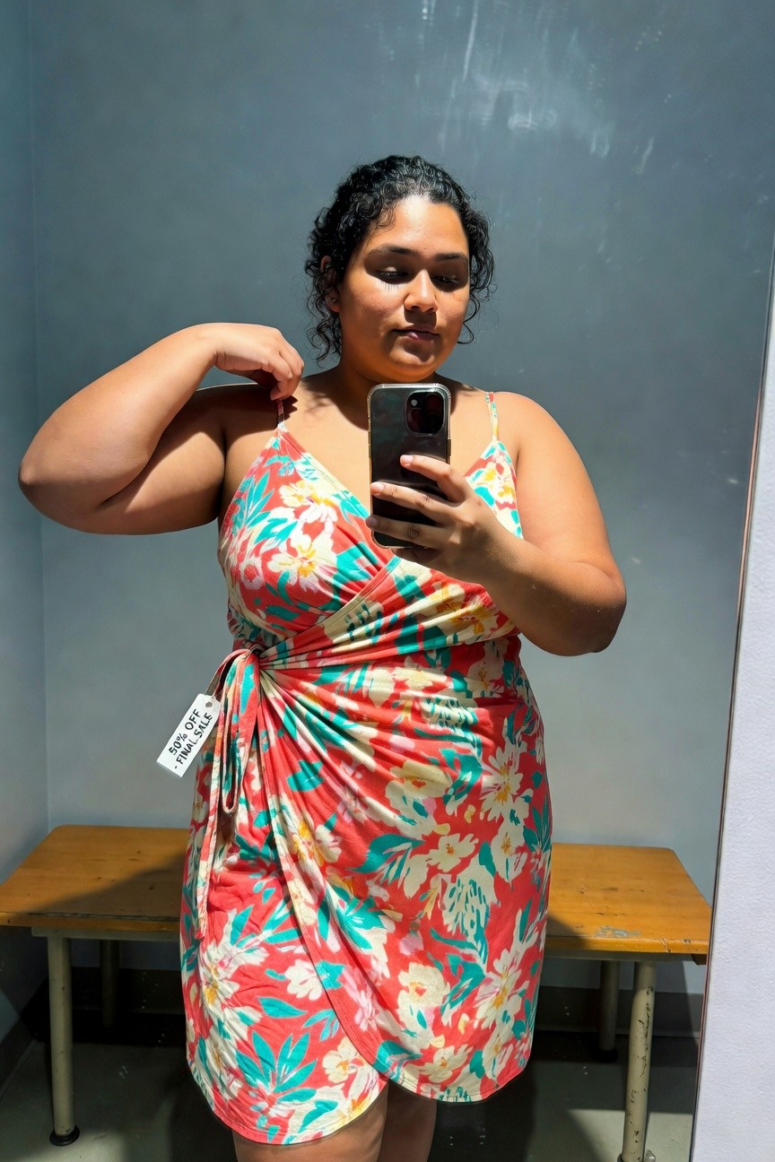 Plus-size woman with curly dark hair taking a mirror selfie in a dressing room, wearing a bright floral wrap dress in orange, pink, and green hibiscus print with spaghetti straps, holding her phone, price tag visible on the side tie, wooden bench in background.