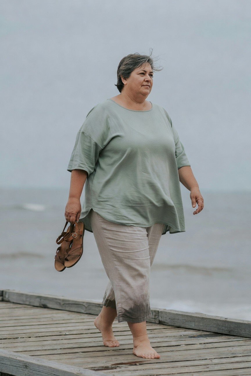 Plus-size woman with short gray hair walking barefoot on a wooden pier, wearing an oversized mint green short-sleeve top, cropped beige linen pants, and holding tan leather sandals in one hand