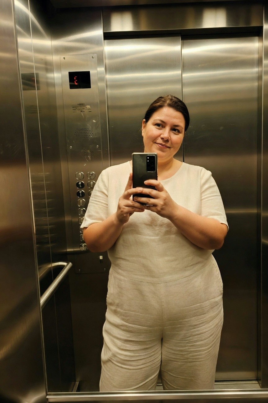 Plus-size woman with brown hair smiling in elevator mirror selfie, wearing short-sleeve white linen wide-leg jumpsuit, holding phone
