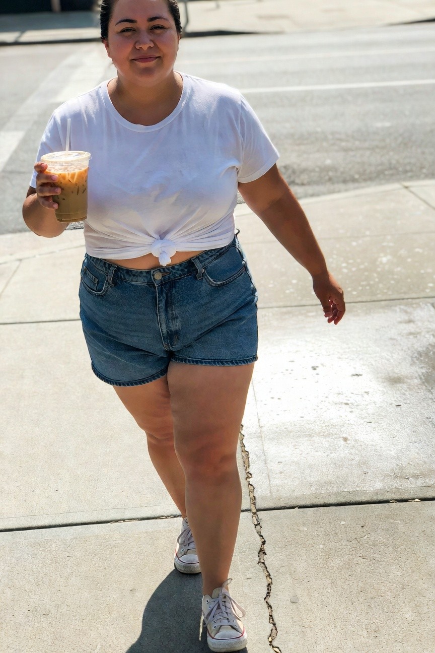 Plus-size woman walking in a knotted white t-shirt, high-waisted cuffed blue denim shorts, and white sneakers, holding an iced coffee