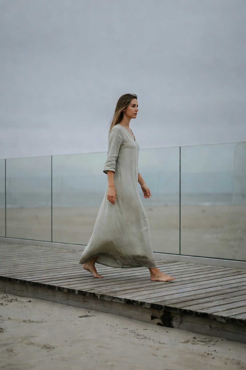 Side view of a woman walking barefoot in a loose sage green linen maxi dress with V-neck and three-quarter sleeves, on a wooden pier with glass railing and beach in background