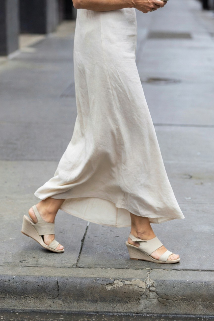 Side view of a woman walking in a flowing beige maxi dress with nude strappy wedge sandals, holding a phone, on a city sidewalk
