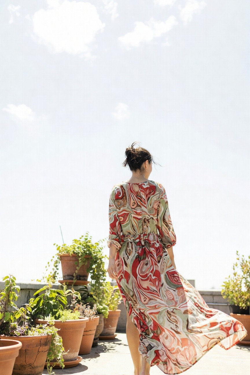 Woman viewed from behind wearing a flowing long-sleeved maxi dress with bold swirling red, green, and cream paisley print, standing barefoot on a terrace surrounded by potted plants