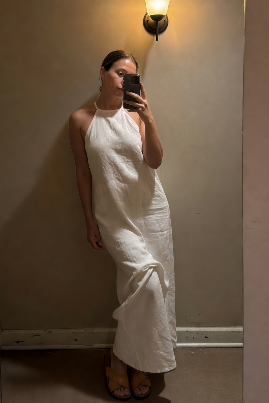Woman taking a mirror selfie in a white linen halter neck maxi dress with a flowing skirt, tan crossed-strap sandals, simple hoop earrings, leaning against a beige wall indoors