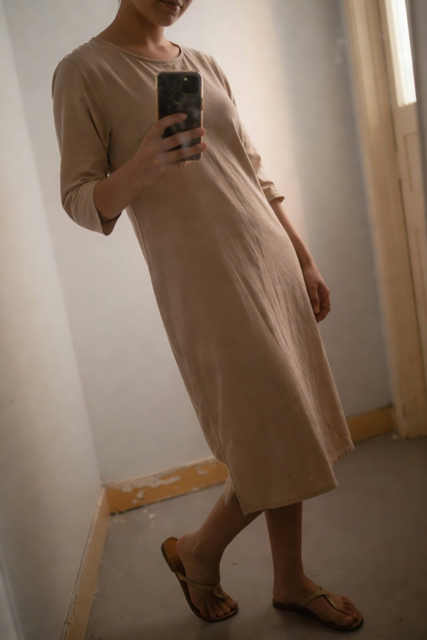Woman in a beige long-sleeved midi dress with side slit, standing sideways holding a smartphone for a selfie, wearing tan flat sandals, indoor setting with white walls.