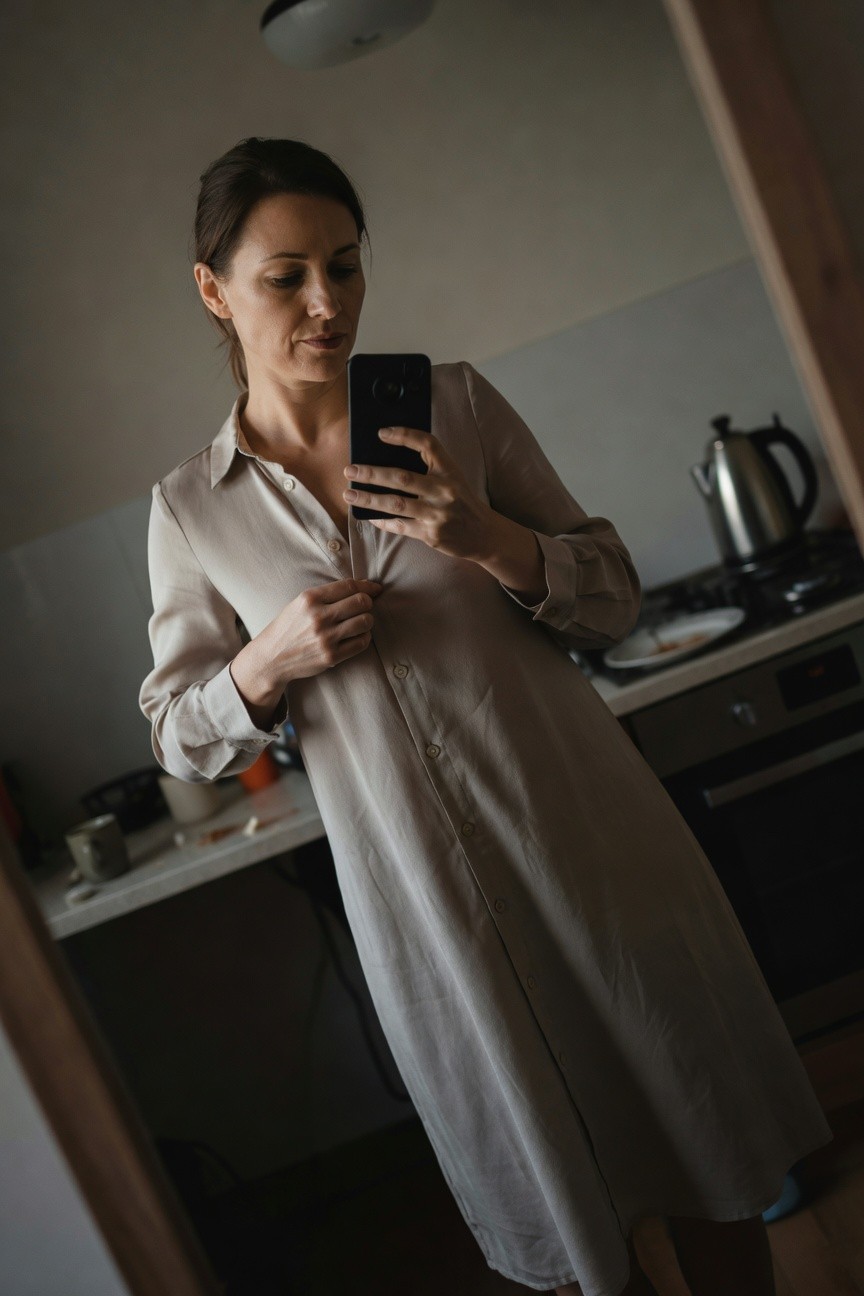 Woman taking a mirror selfie in a beige long-sleeve button-front shirt dress, slightly unbuttoned at the collar, standing in a kitchen with one hand adjusting a button, hair in a ponytail, barefoot on wood floor.
