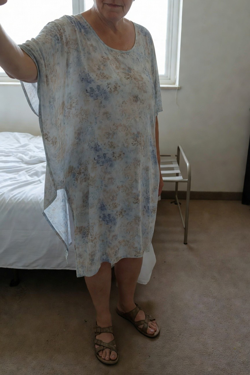 Mature plus-size woman standing in a bedroom wearing a loose light blue and beige floral print dress with open dolman sleeves and mid-length hem, paired with tan cross-strap flat sandals, one arm raised