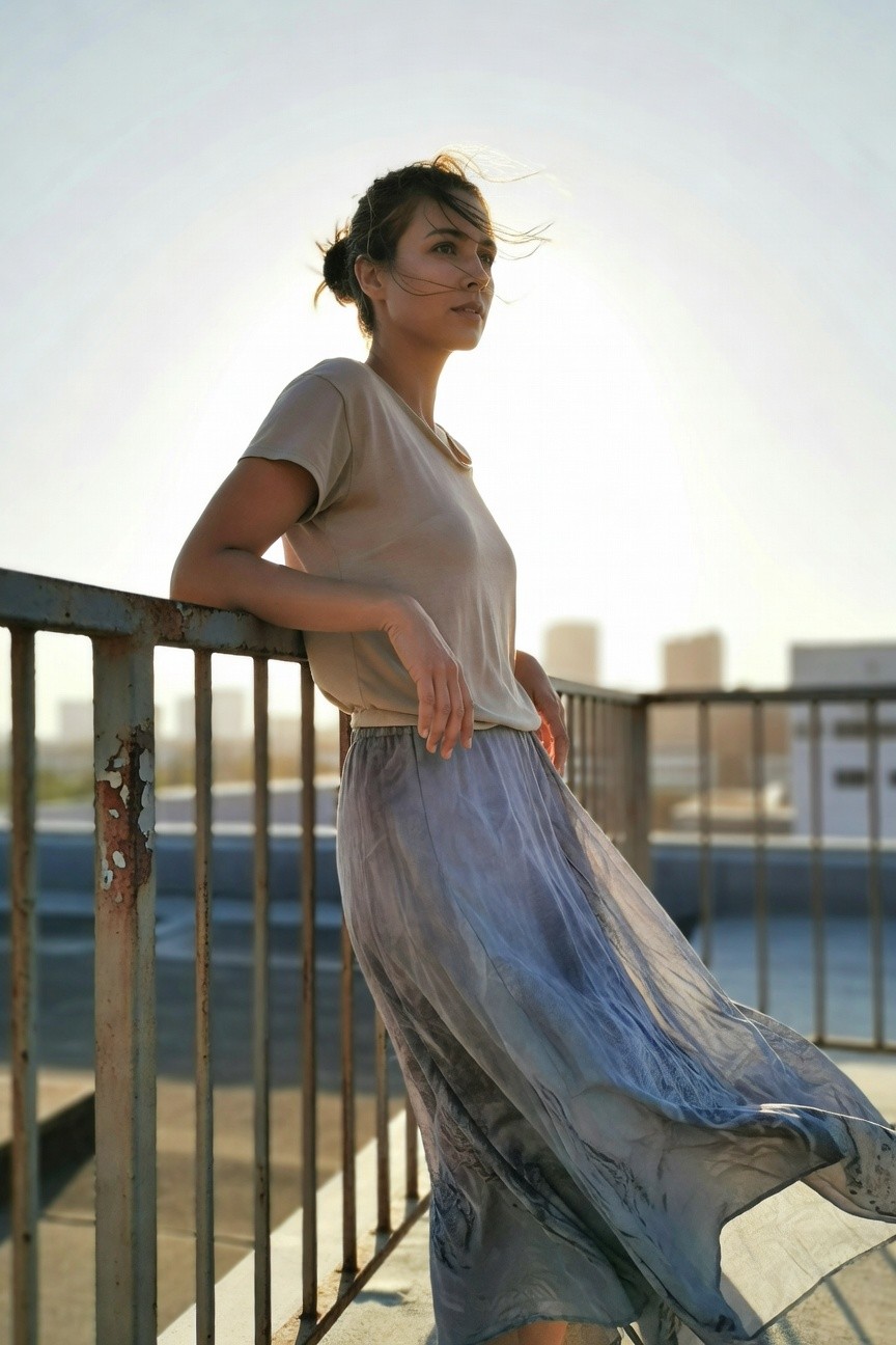 Side view of woman in loose beige short-sleeve t-shirt and flowing gray pleated midi skirt, leaning casually on metal railing
