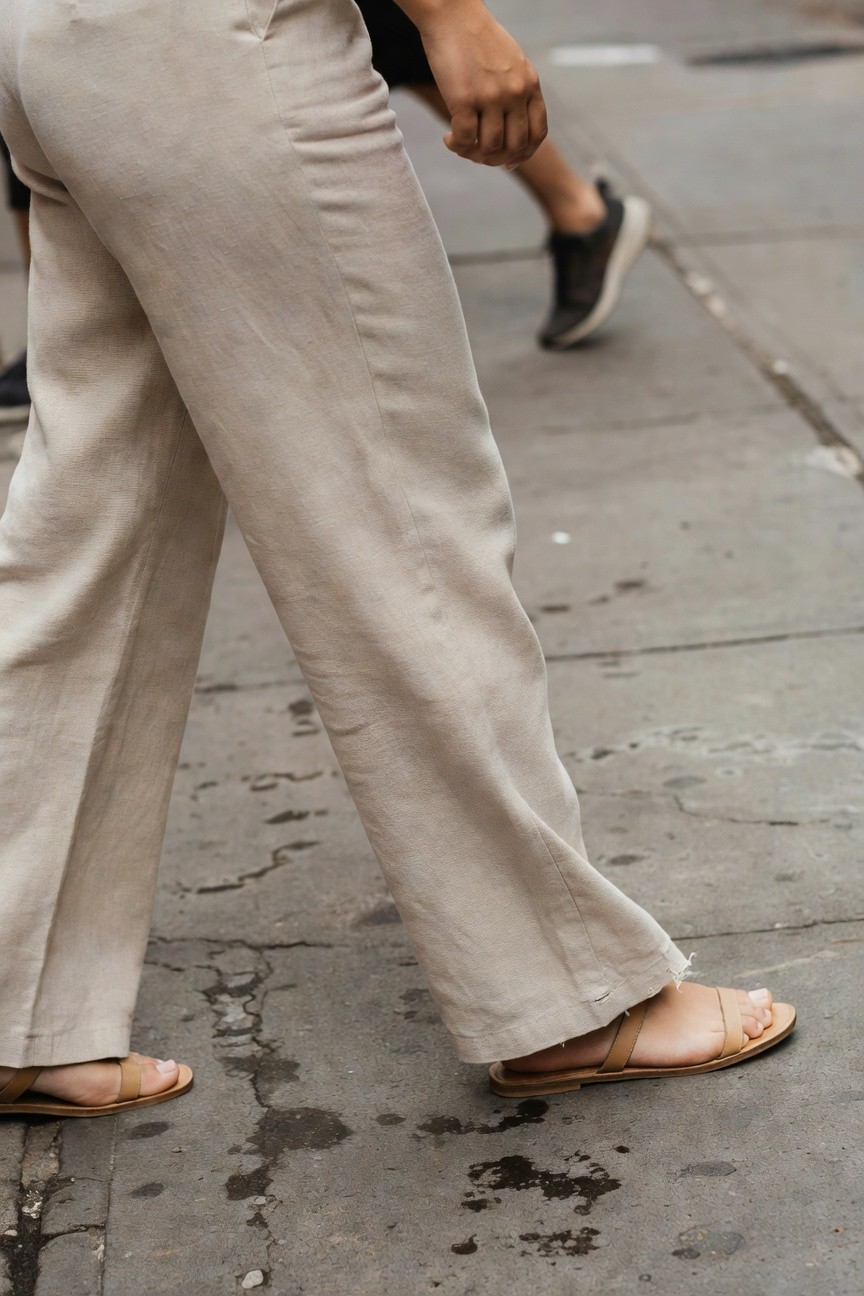 Rear view of a woman walking in light beige wide-leg linen pants and tan flat strappy sandals on a paved sidewalk