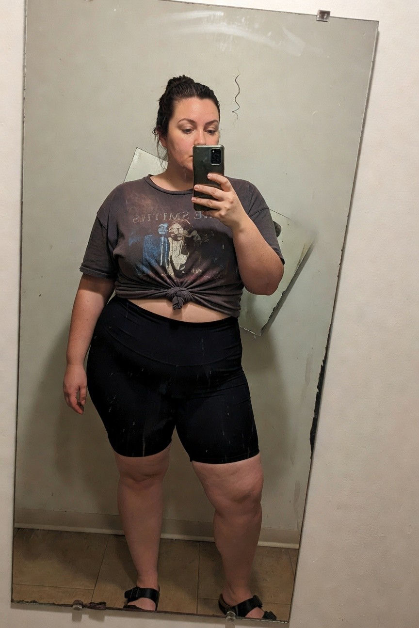 Plus-size woman taking a mirror selfie in a bathroom, wearing a knotted oversized gray Smiths band T-shirt, high-waisted black biker shorts, and black strappy flat sandals, standing confidently with phone in hand