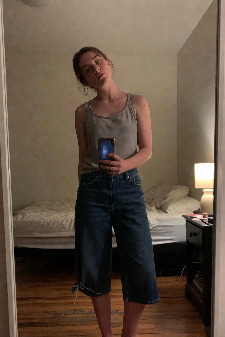 Petite woman taking a mirror selfie in a bedroom, wearing a loose gray sleeveless tank top tucked slightly into high-waisted wide-leg cropped blue jeans with frayed tied hems, standing barefoot on wooden floor