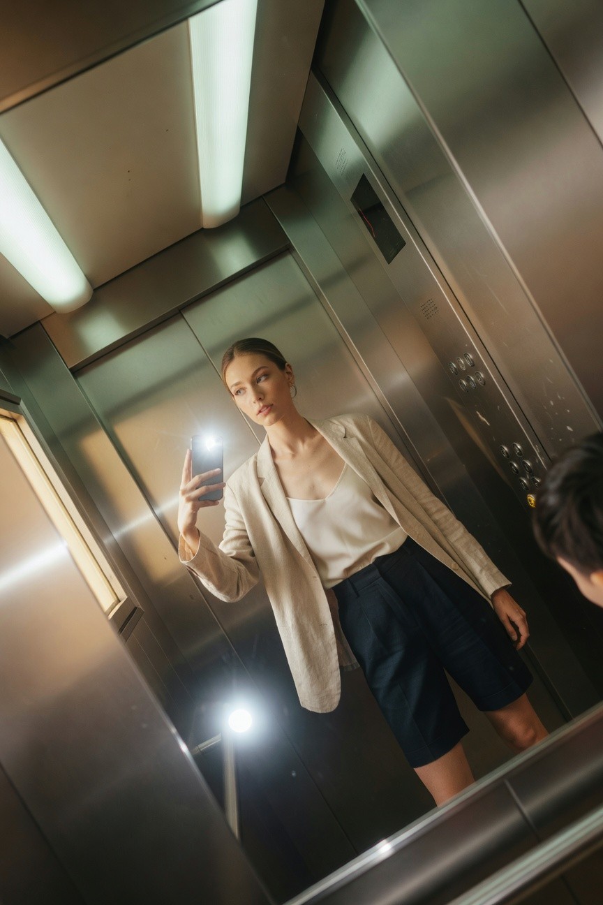 Petite woman taking a mirror selfie in an elevator wearing a light cream linen blazer over white silk camisole and navy tailored shorts with gold hoop earrings