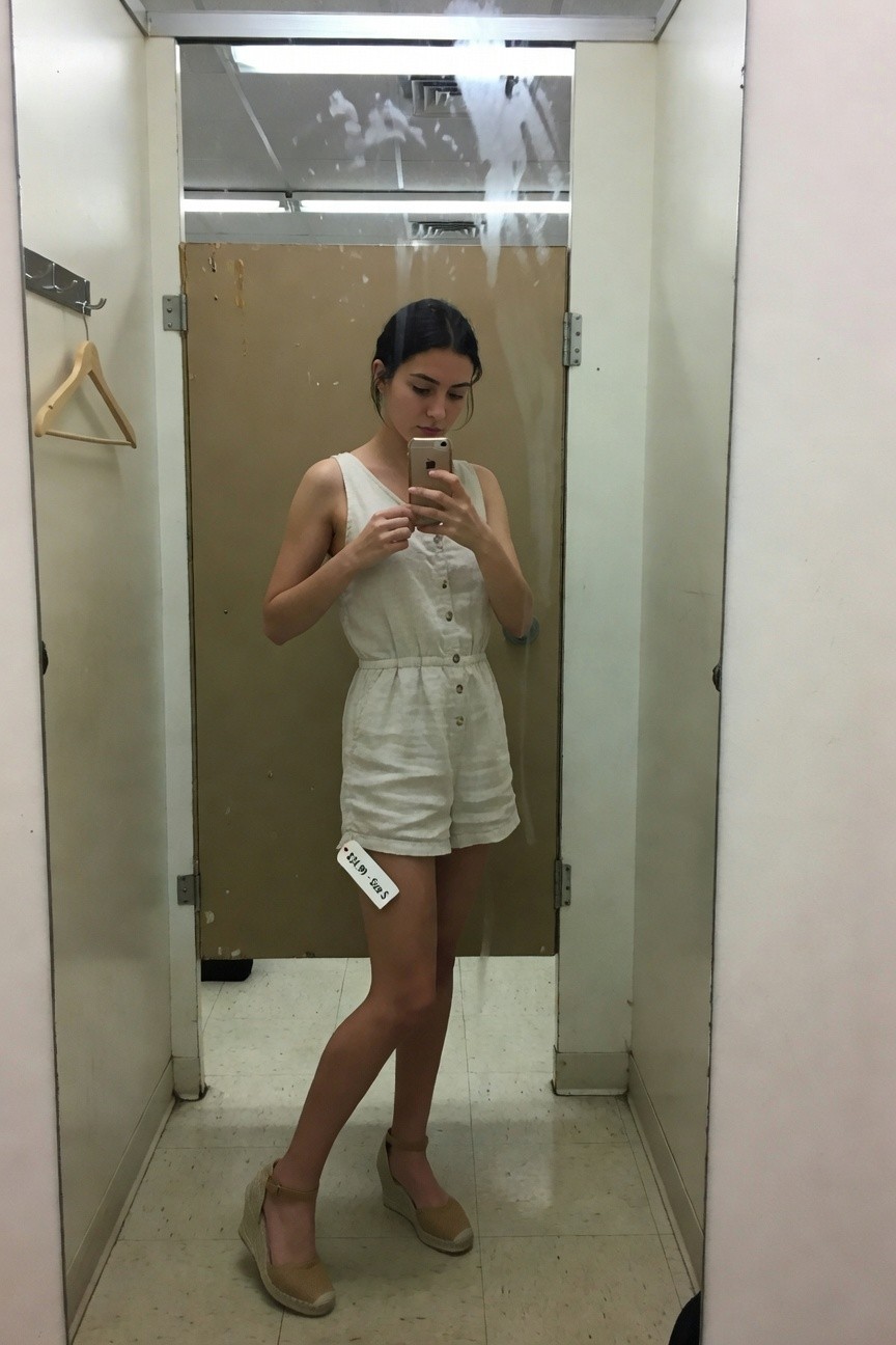 Petite woman taking a mirror selfie in a store fitting room, wearing a white sleeveless linen romper with button front and waist tie, short inseam, tan espadrille wedge sandals, price tag visible on thigh