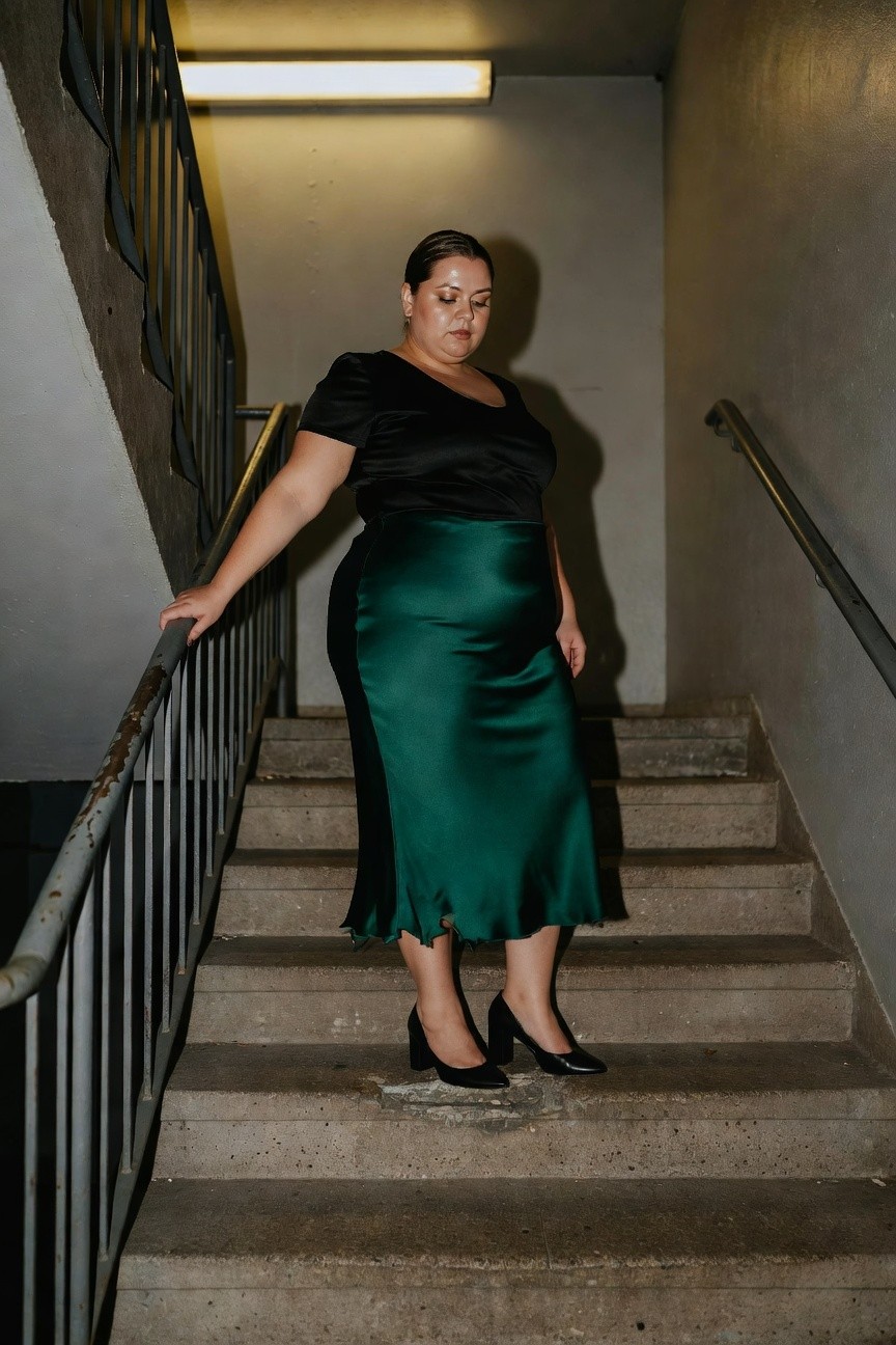 Plus size woman standing on concrete stairs in a black short-sleeved wrap top, emerald green satin flared midi skirt, and black mid-heel pumps, hand on metal railing in dimly lit stairwell