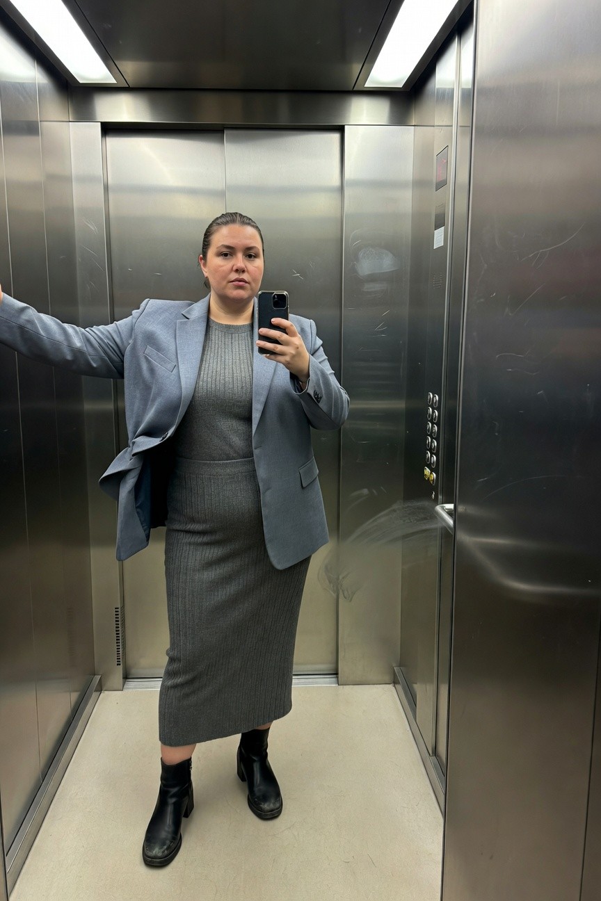 Plus-size woman in an open blue blazer over a gray ribbed knit midi dress with black block-heel ankle boots, posing with arms outstretched in a silver elevator for a selfie