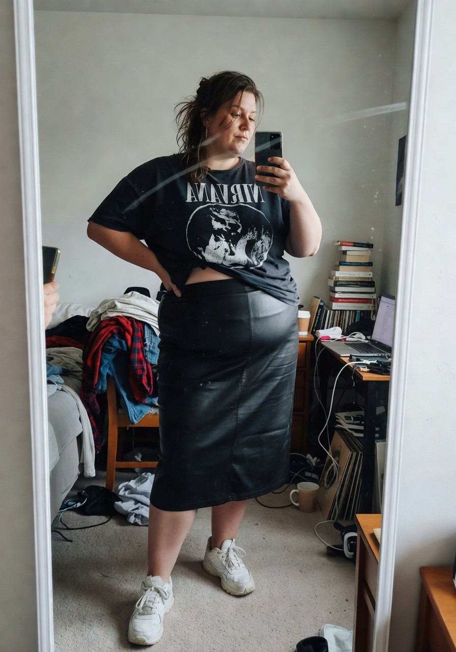 Plus-size woman in oversized black graphic tee reading The Neon Owls tucked slightly over high-waisted black leather midi skirt and white chunky sneakers posing confidently hands on hips in bedroom mirror selfie