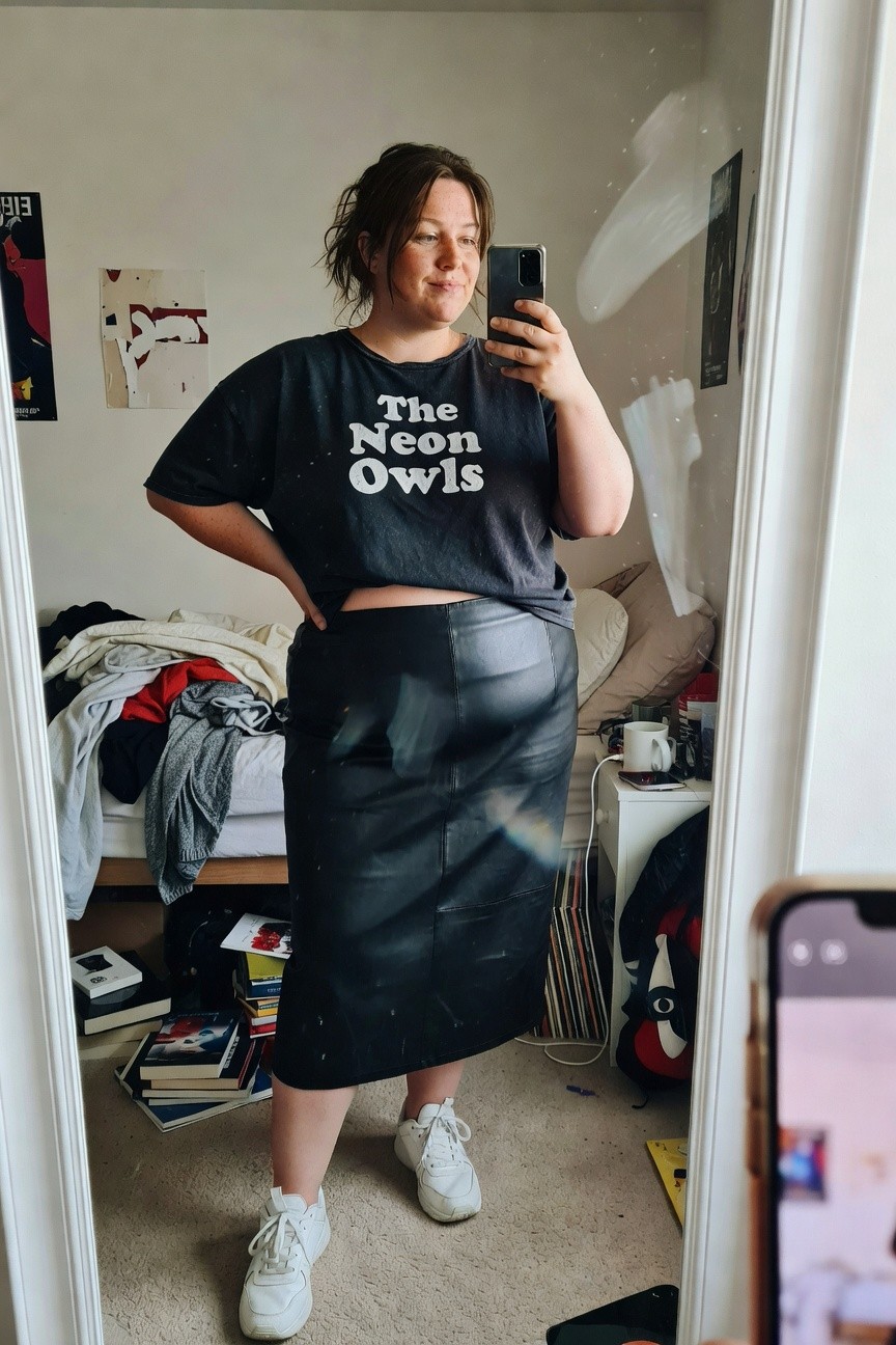 Plus-size woman in oversized black graphic tee reading The Neon Owls tucked slightly over high-waisted black leather midi skirt and white chunky sneakers posing confidently hands on hips in bedroom mirror selfie