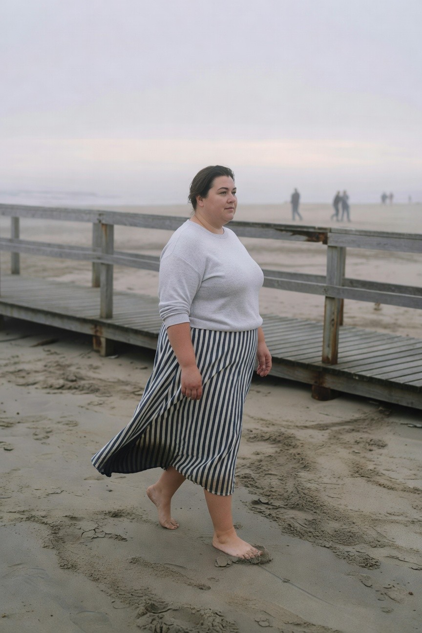 Plus-size woman walking barefoot on sand near a wooden boardwalk, wearing a light grey oversized sweater and navy-and-white striped midi skirt that flows as she moves, side profile view.