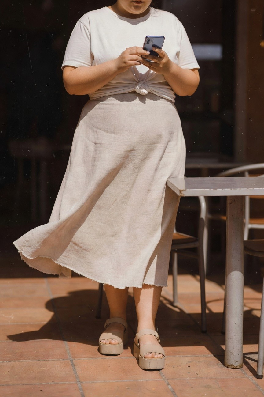 Plus-size woman standing outdoors near a metal cafe table, wearing a white short-sleeve t-shirt knotted at the waist, flowy beige linen midi skirt with fringed hem, and beige platform sandals while holding a smartphone