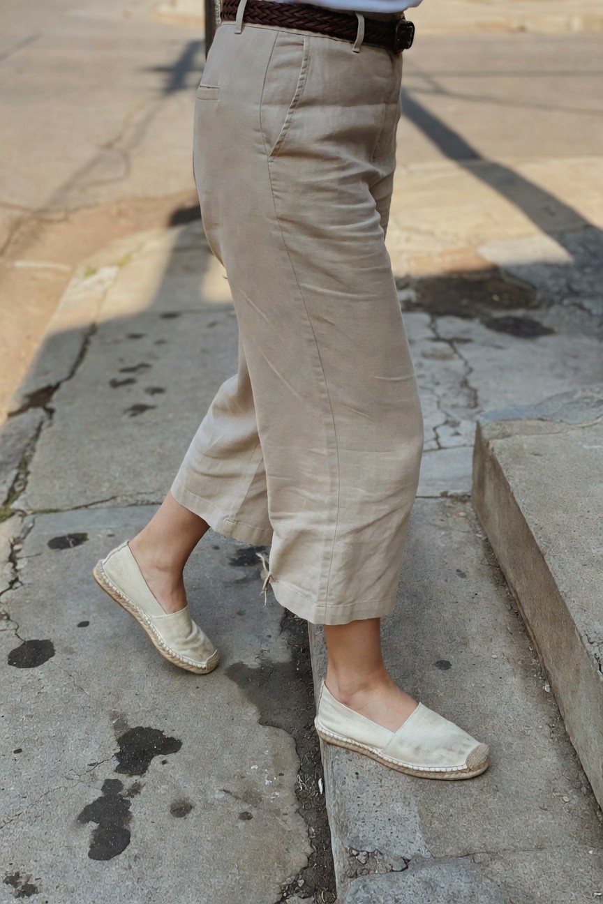 Woman in beige wide-leg cropped linen pants with rolled cuffs, brown braided leather belt, white tucked top, and white canvas espadrilles with jute soles, walking up concrete steps