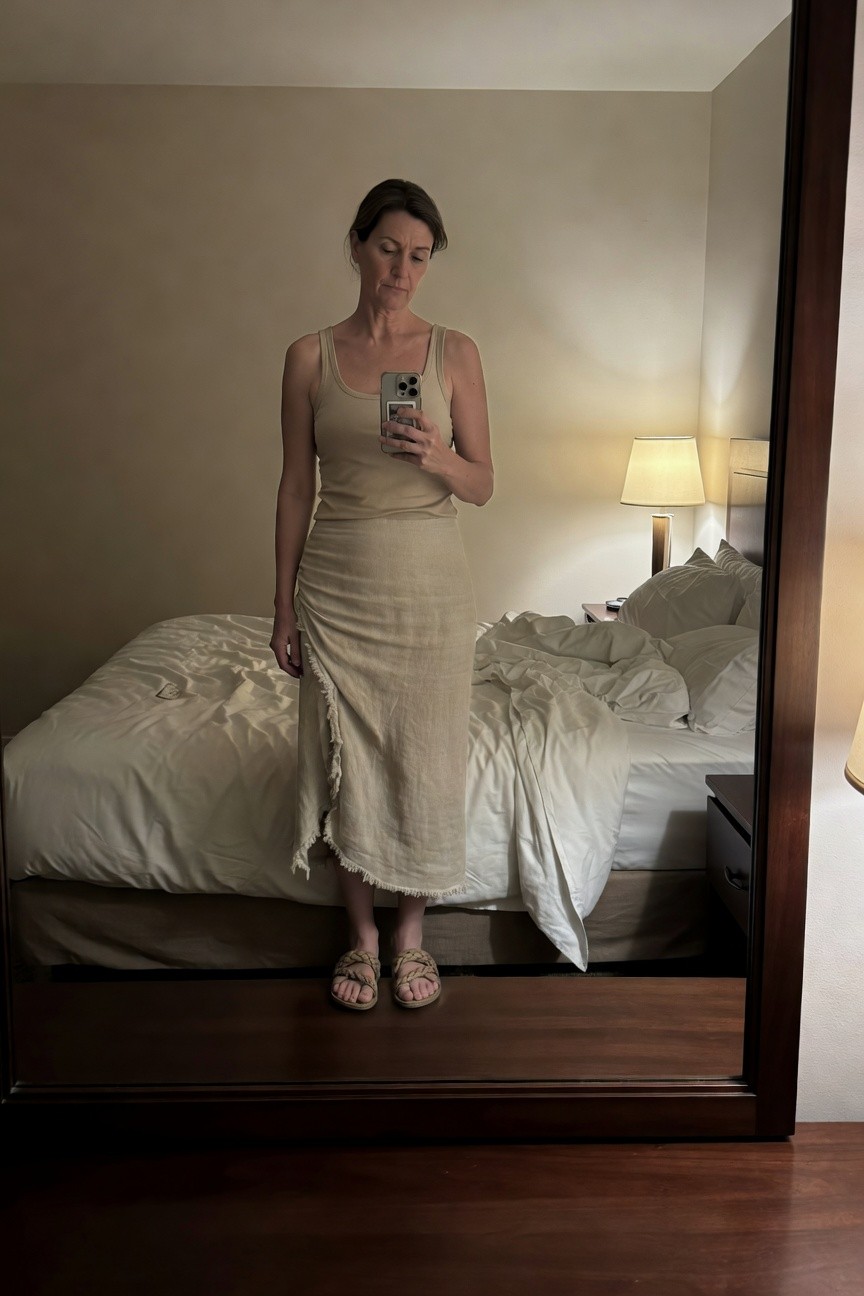 Woman in beige spaghetti-strap tank top and matching fringed-hem linen midi skirt standing in front of full-length mirror, wearing tan braided flat sandals, neutral hotel room interior visible in reflection