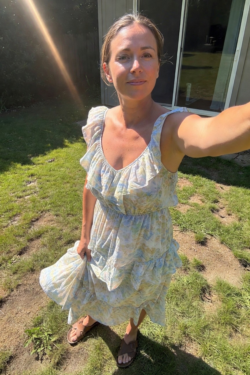Woman selfie in a light floral print tiered maxi dress with ruffled shoulders and spaghetti straps, tan strappy sandals, holding skirt in backyard grass