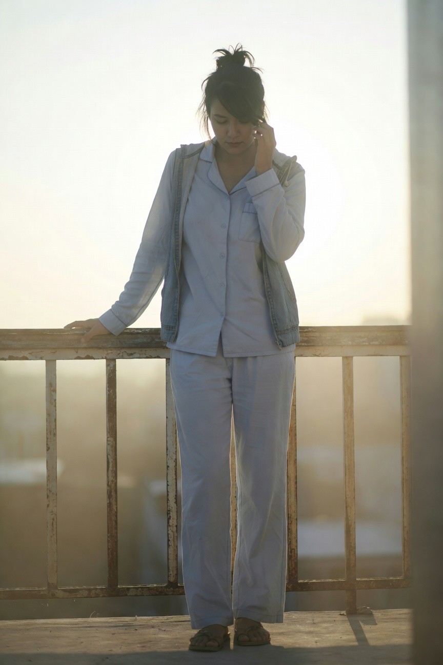 Woman leaning on balcony railing in pale blue long-sleeve button-up shirt, matching straight-leg pants, open light-wash denim vest, phone to ear, messy bun hairstyle, wearing tan flip-flop sandals