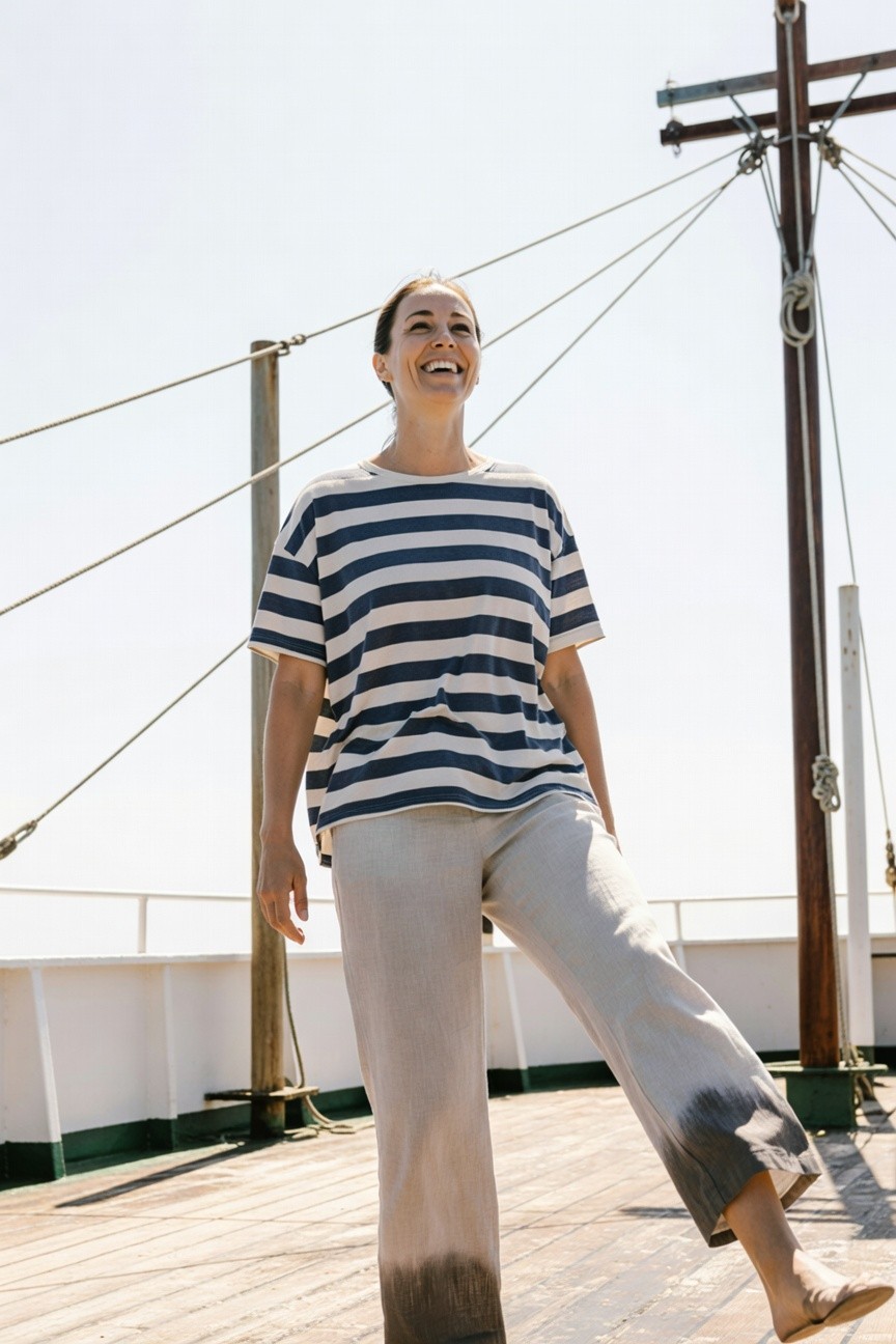 Woman smiling on boat deck in oversized navy-and-white striped short-sleeve tee, wide-leg light beige linen pants fading to darker hems, and tan flat sandals