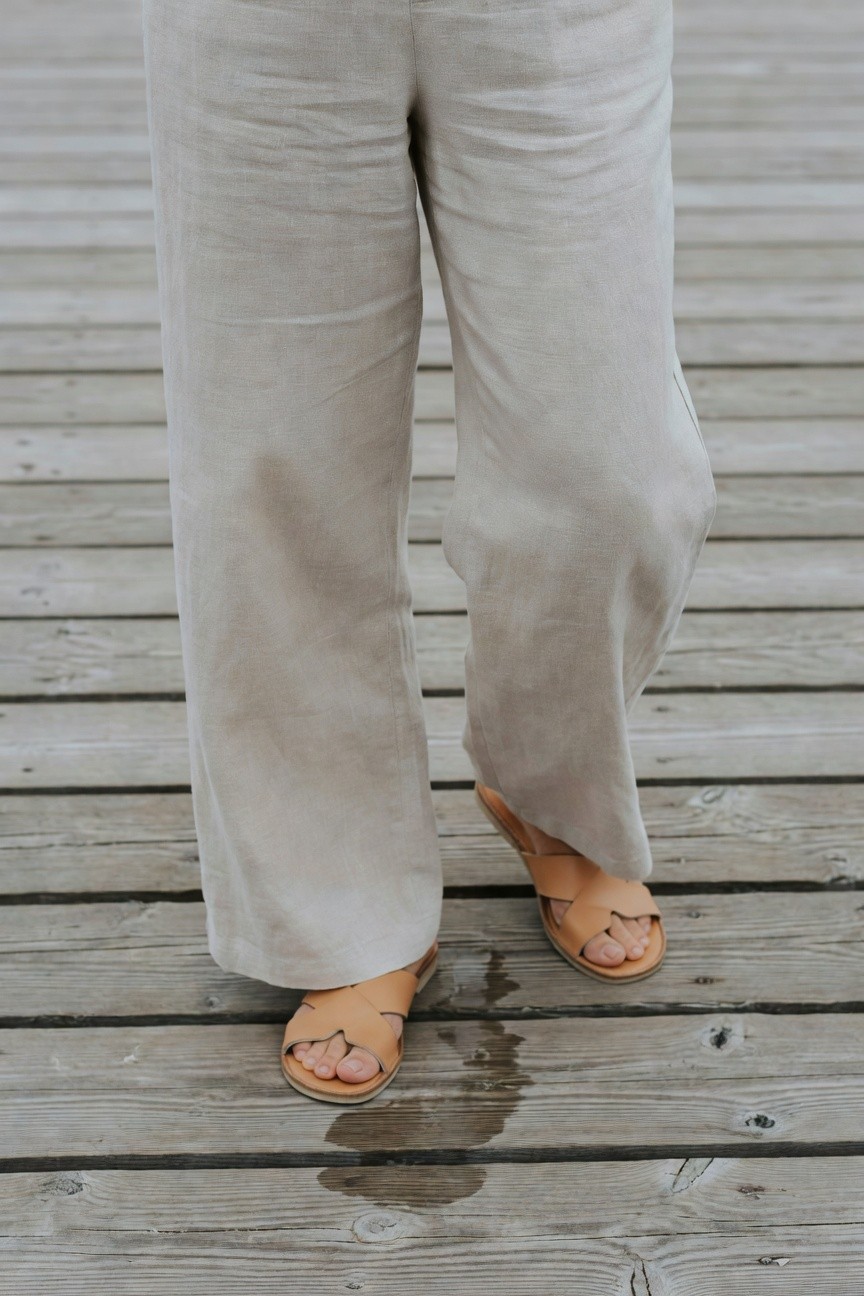 Woman's lower body in wide-leg light beige linen pants tucked slightly at ankles over tan leather cross-strap sandals on wooden surface with water spots nearby