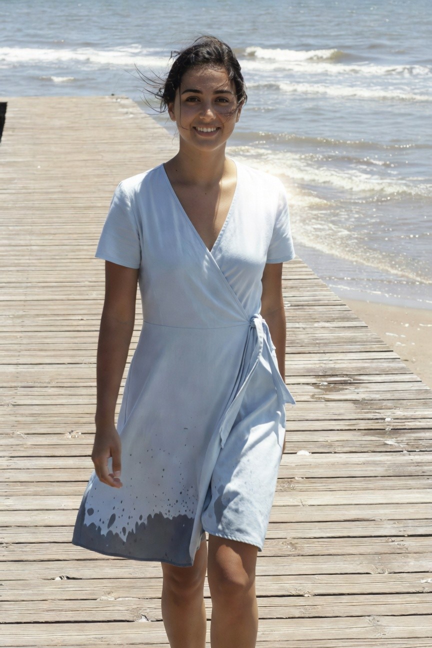 Young woman with dark wavy hair smiling while walking on a wooden pier, wearing a short-sleeved light blue wrap dress with white and gray splatter fading into darker blue at the hem, V-neckline, side tie, against ocean backdrop