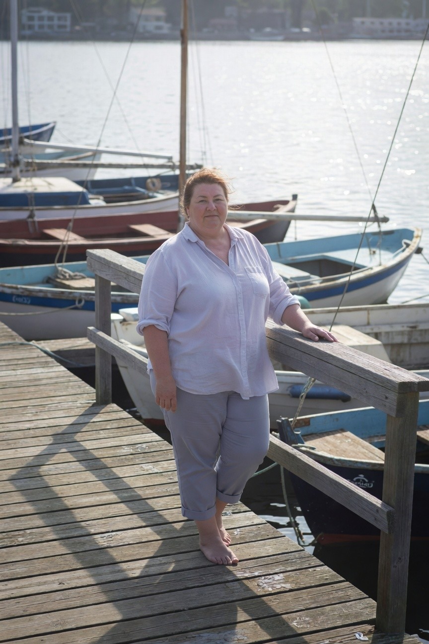 Plus-size woman standing on a wooden dock by a lake, wearing a loose light gray-white button-up shirt with rolled sleeves and matching light gray wide-leg pants cuffed at the ankles, barefoot, leaning on the railing with boats moored nearby in the background.